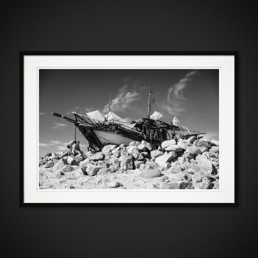The Last Mermaid — A figure lies across a wrecked boat on a rocky shore. Framing preview by Albert Pocej; frame not included.